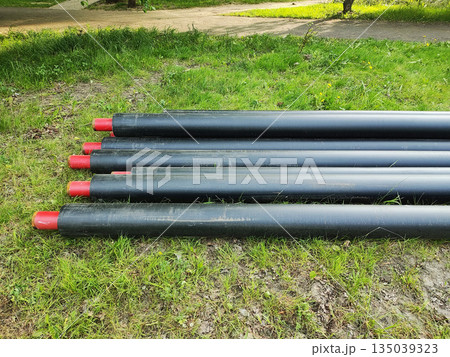 Row of large black pipes with red protective caps laid horizontally on green grass beside paved path under natural light, outdoor scene, no people. 135039323
