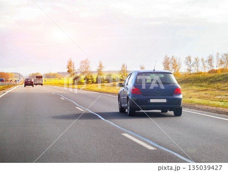 Blue hatchback car driving on highway during autumn day. Road trip scene with vehicle traveling at speed. Transportation concept with bright sun flare. 135039427
