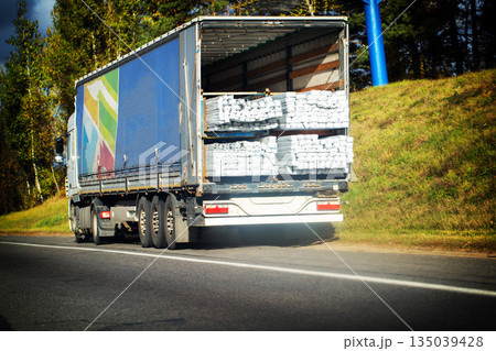 Heavy duty semi-trailer truck with open rear of trailer revealing cargo of white bags, vehicle driving on asphalt road with green trees and grassy embankment under sky. 135039428