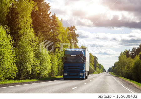 Modern truck transports cargo against the backdrop of forest and sun in summer, beautiful sky with clouds. Logistics company delivering cargo on international routes. Copy space for text. Freight Modern truck transports cargo against the backdrop of forest and sun in summer, beautiful sky with clouds. Logistics company delivering cargo on international routes. Copy space for text. Freight 135039432