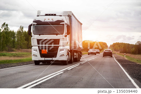 White semi truck with cargo trailer traveling on rural highway against sunset sky, transportation of goods and logistics business White semi truck with cargo trailer traveling on rural highway against sunset sky, transportation of goods and logistics business 135039440