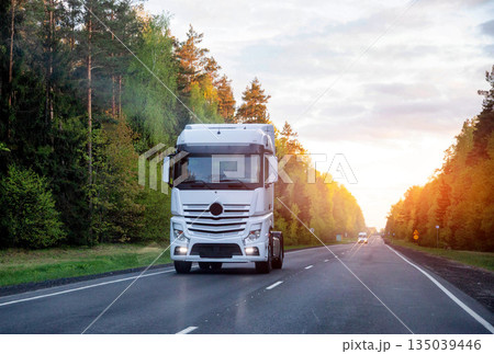 White semi-trailer truck drives on paved rural road through dense forest. Sunlight illuminates trees during sunset. Essential freight transportation. White semi-trailer truck drives on paved rural road through dense forest. Sunlight illuminates trees during sunset. Essential freight transportation. 135039446