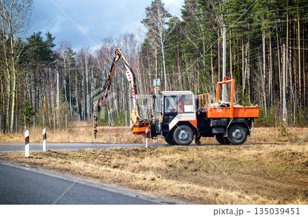 An orange and white utility vehicle with a drilling boom is parked on the side of a paved road during daylight hours. Road maintenance and pole installation. 135039451