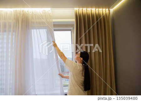 Woman hand adjusting sheer white curtain, revealing bright daylight through window, cozy indoor setting with warm ambient light, comfort feeling 135039480