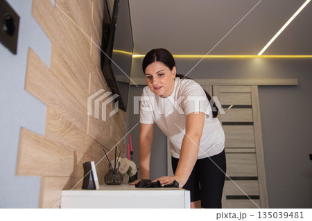 Woman wiping dust from surface of white furniture in modern apartment living room, home cleaning routine, domestic chore, daily dwelling maintenance. 135039481