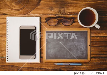 Showing flat-design overhead desk with pad and smartphone, framed chalkboard, glasses, coffee, pen Showing flat-design overhead desk with pad and smartphone, framed chalkboard, glasses, coffee, pen 135039871
