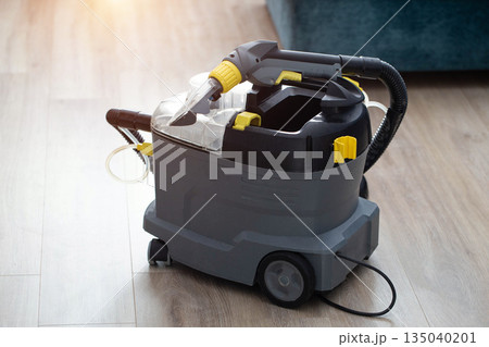 A modern upholstery vacuum cleaner stands in a room against a sunset backdrop. Cleaning service, industry A modern upholstery vacuum cleaner stands in a room against a sunset backdrop. Cleaning service, industry 135040201