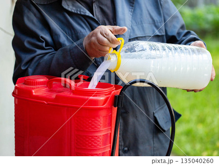 Close up view of gardener preparing solution for plant treatment, pouring liquid from plastic bottle into red agricultural sprayer for parasite and disease protection Close up view of gardener preparing solution for plant treatment, pouring liquid from plastic bottle into red agricultural sprayer for parasite and disease protection 135040260