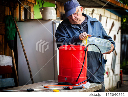 Elderly man prepares solution for plant pest control, pouring liquid from plastic bottle into red garden sprayer for dacha agriculture work Elderly man prepares solution for plant pest control, pouring liquid from plastic bottle into red garden sprayer for dacha agriculture work 135040261