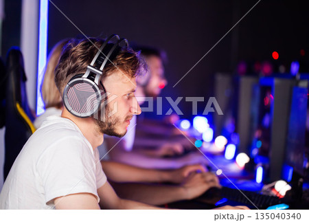 Young man playing video game, wearing headphones, in cyber club, intense focus of player, e-sports competition of gamers, internet cafe tournament 135040340