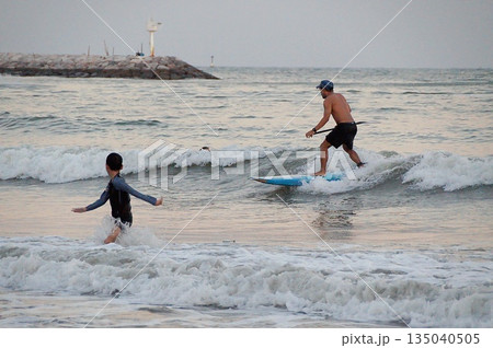 A young surfer rides a wave on a wooden board in the blue sea. A child plays in the waves nearby.Young surfer and child. A young surfer rides a wave on a wooden board in the blue sea. A child plays in the waves nearby.Young surfer and child. 135040505