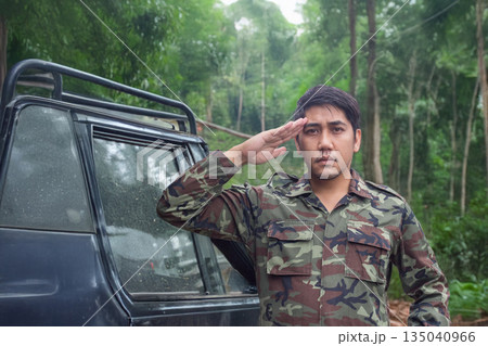 An Asian army officer in uniform salutes directly at the camera, deep in a lush jungle with an off-road vehicle visible in the background An Asian army officer in uniform salutes directly at the camera, deep in a lush jungle with an off-road vehicle visible in the background 135040966
