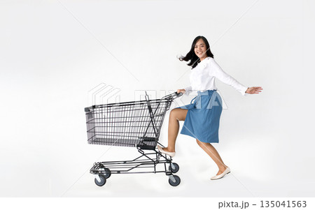 Joyful Asian woman playfully pushing a shopping cart on a clean white background, perfect for advertising and marketing campaigns. Captures consumer happiness and effortless retail experiences 135041563