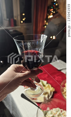 Hand holding glass of red wine above festive table with food and blurred christmas lights in background. This image represents celebration holiday atmosphere and cozy home gathering moment Hand holding glass of red wine above festive table with food and blurred christmas lights in background. This image represents celebration holiday atmosphere and cozy home gathering moment 135041939