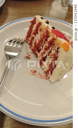 Slice of red velvet cake with cream frosting served on plate with fork on wooden table. This dessert image represents indulgence comfort food and celebration moment Slice of red velvet cake with cream frosting served on plate with fork on wooden table. This dessert image represents indulgence comfort food and celebration moment 135041946