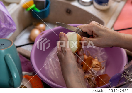 Close-Up Of Hand Peeling An Onion With Knife Over Purple Bowl In The Kitchen 135042422