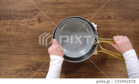 Hands hold a gray nonstick frying pan with golden handles above a wooden tabletop. The clean background and composition focus on the cookware details and form. Hands hold a gray nonstick frying pan with golden handles above a wooden tabletop. The clean background and composition focus on the cookware details and form. 135042878