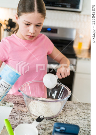 Young girl wearing a pink shirt measures baking powder from a container into a clear bowl. The granite counter holds other kitchen tools and utensils. 135042879