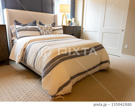 Modern bedroom interior with a neatly made bed featuring striped bedding and accent pillows. Matching bedside lamps provide warm lighting, creating a cozy and inviting mood. The space has a balanced 135042880
