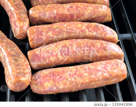 A row of raw sausages is arranged side by side on a black grill. The meat is pink and marbled, ready to begin the cooking process. A row of raw sausages is arranged side by side on a black grill. The meat is pink and marbled, ready to begin the cooking process. 135042896