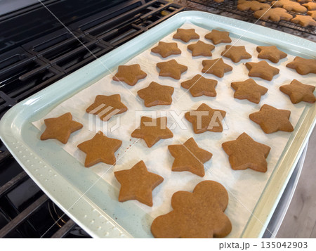 A baking tray lined with parchment paper holds freshly baked gingerbread cookies in star and gingerbread-person shapes. The tray rests on a stovetop with warm kitchen lighting revealing their golden A baking tray lined with parchment paper holds freshly baked gingerbread cookies in star and gingerbread-person shapes. The tray rests on a stovetop with warm kitchen lighting revealing their golden 135042903