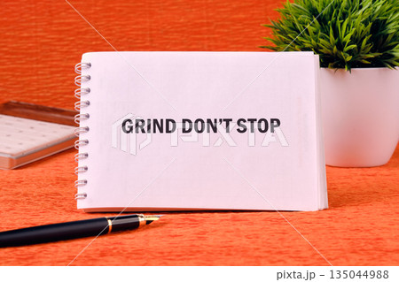 A white notepad shows the text GRIND DONT STOP, placed on an orange surface with a pen and potted plant beside it, symbolizing perseverance and hard work A white notepad shows the text GRIND DONT STOP, placed on an orange surface with a pen and potted plant beside it, symbolizing perseverance and hard work 135044988