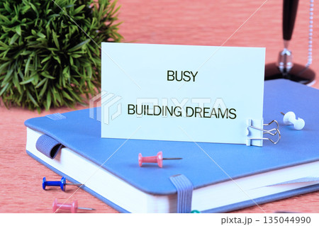 A serene scene presents a notebook with a motivational note card perched upon it, complemented by stationery like pushpins and a binder clip. A verdant plant adds a touch of nature 135044990