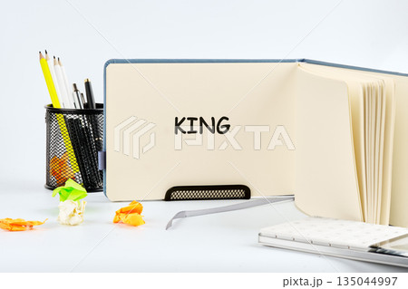 An open notebook displays the word King. Pencils, pens, and crumpled papers scatter around the desk near a book stand. A journal lays near the notebook An open notebook displays the word King. Pencils, pens, and crumpled papers scatter around the desk near a book stand. A journal lays near the notebook 135044997