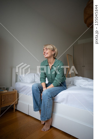 Carefree positive woman sitting on bed in bedroom and smiling brightly, showing self love and peace. 135045844