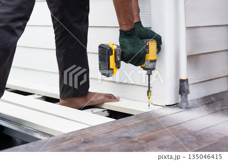 A worker is using a cordless power drill to fasten exterior floor boards. 135046415