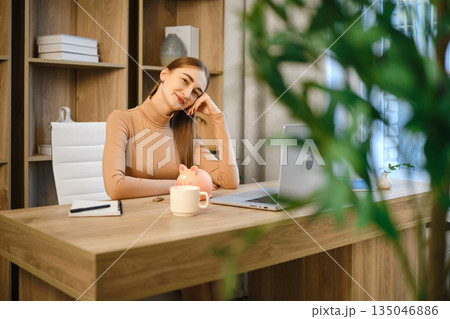 Woman sitting at desk with laptop, mug, and piggy bank in a modern office space 135046886