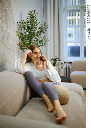 Woman sitting on a couch talking on the phone in a bright room with plants 135046901
