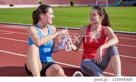 Two smiling young women in athletic wear resting on a red running track at a sunny stadium. They are holding water bottles, talking, and laughing after a successful training session. Healthy lifestyle 135046986