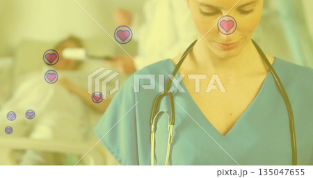 Standing nurse in scrubs, stethoscope bowing head at hospital bedside, floating hearts, copy space 135047655