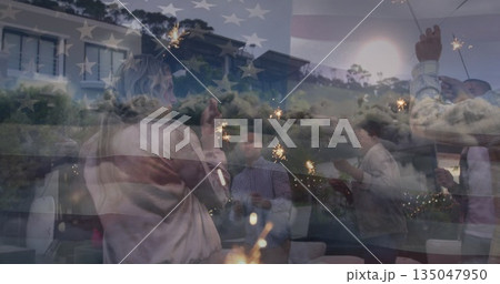 Six friends holding sparklers beside patio chairs and low coffee tables at dusk, with string lights 135047950