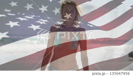 Walking woman in bikini along sandy shoreline with lapping ocean waves, with American flag overlay 135047956