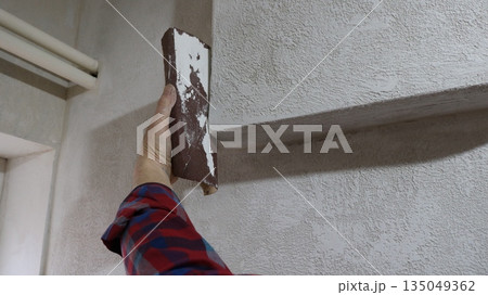 A homemade hand tool for peeling and polishing walls, a piece of sandpaper in the hand of a worker sharpening a convex part of a wall, an emery board in the process of leveling plaster on a wall A homemade hand tool for peeling and polishing walls, a piece of sandpaper in the hand of a worker sharpening a convex part of a wall, an emery board in the process of leveling plaster on a wall 135049362