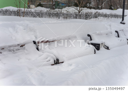 Snowy winter street. Snow covered park benches surrounded by deep snowdrifts during heavy snowfall. Concept of winter abandonment, seasonal challenges, frozen cityscape Snowy winter street. Snow covered park benches surrounded by deep snowdrifts during heavy snowfall. Concept of winter abandonment, seasonal challenges, frozen cityscape 135049497