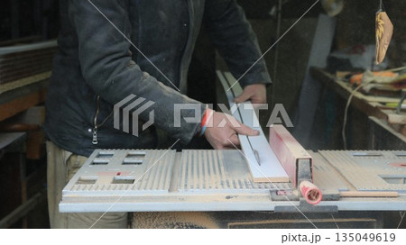 An impersonal carpenter works on a circular saw, cutting a narrow wooden plank lengthwise against the background of an equipped workshop, machine processing and preparation of panels for construction An impersonal carpenter works on a circular saw, cutting a narrow wooden plank lengthwise against the background of an equipped workshop, machine processing and preparation of panels for construction 135049619