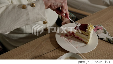 Older woman savoring delicious layered cake. Close-up shot of hand holding fork. Creamy dessert with berry topping on plate. Cozy and relaxed atmosphere. 135050194