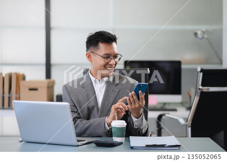 Businessman using laptop computer and smartphone in office. Happy man, entrepreneur, small business owner working online. Businessman using laptop computer and smartphone in office. Happy man, entrepreneur, small business owner working online. 135052065