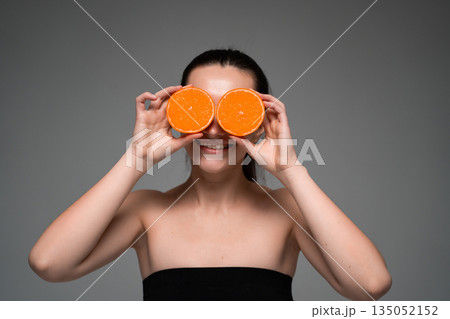Woman holding fresh orange halves over eyes on neutral background, creative healthy lifestyle portrait with citrus fruit, vitamins, wellness and natural beauty 135052152