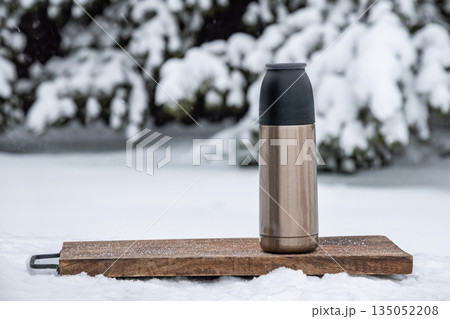 Metal thermos standing on wooden serving board in snowy outdoor landscape. Concept of hot drink, travel, winter picnic and outdoor warmth. 135052208