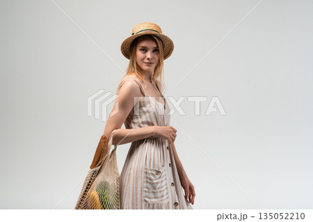 Young woman walking with reusable mesh shopping bag filled with groceries, wearing summer dress and straw hat, sustainable lifestyle concept and zero waste shopping 135052210