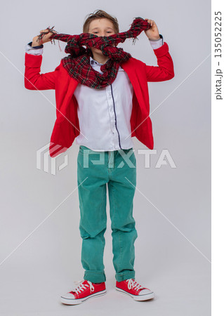 Funny boy in a red jacket, green jeans and sneakers in full growth on a gray background. He hid his face behind a long scarf. 135052225