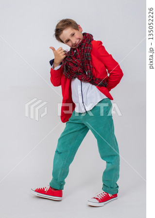 Funny boy in red jacket, green jeans and sneakers in full growth on gray background. He shows sign of class. Funny boy in red jacket, green jeans and sneakers in full growth on gray background. He shows sign of class. 135052226