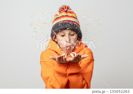 A boy in an orange hoodie and striped hat blows colorful confetti from his hands. A boy in an orange hoodie and striped hat blows colorful confetti from his hands. 135052263