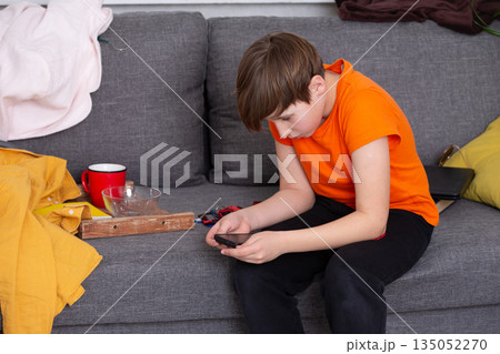 Boy sitting on a couch playing with a smartphone while surrounded by colorful blankets and a mug Boy sitting on a couch playing with a smartphone while surrounded by colorful blankets and a mug 135052270
