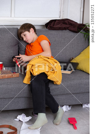 Boy relaxing on a couch wearing an orange shirt and yellow jacket while playing with a toy in a messy room 135052271