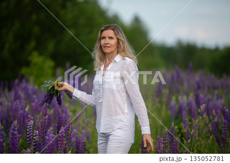 Woman in white outfit walking through vibrant lupine field holding flowers in hand during sunny day Woman in white outfit walking through vibrant lupine field holding flowers in hand during sunny day 135052781